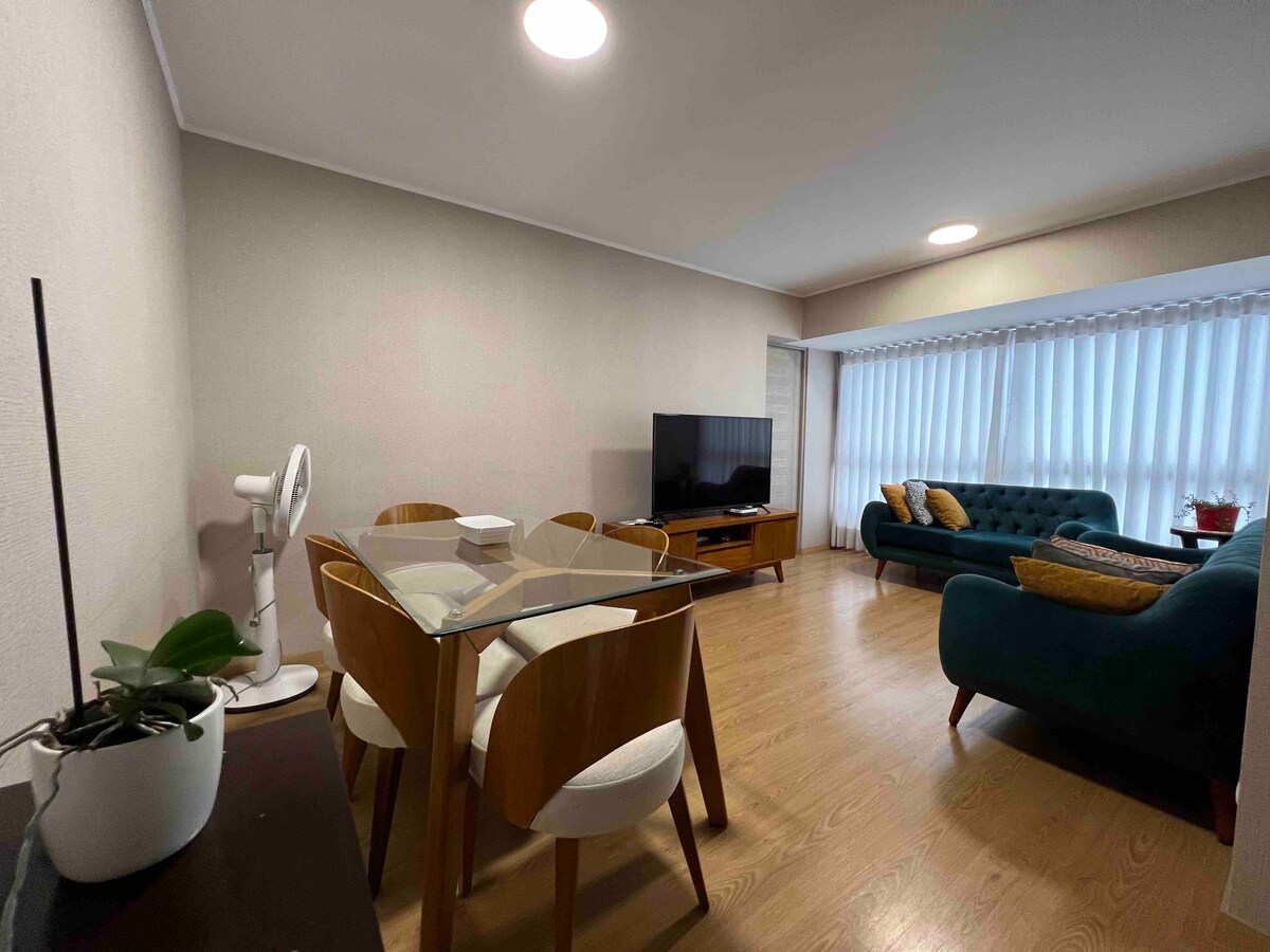 A living and dining area features a glass table surrounded by four wooden chairs. A modern television stands on a stylish wooden console, while a comfortable blue sofa with decorative pillows is positioned nearby. Natural light filters through sheer drapes covering a wide window.