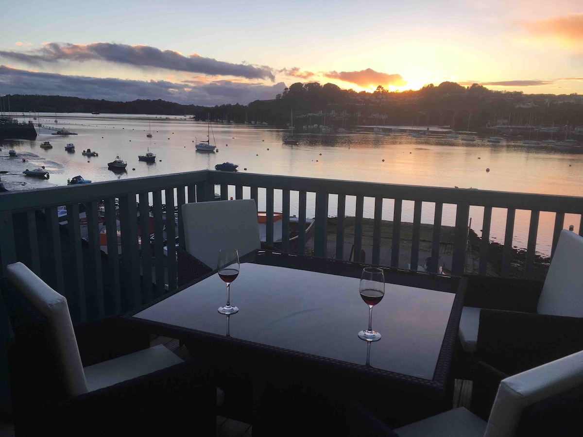 A dining table with two chairs overlooks the tranquil River Tamar at sunset. Two glasses of red wine are placed on the table, reflecting the soft hues of the evening sky, while boats are visible in the serene waters.