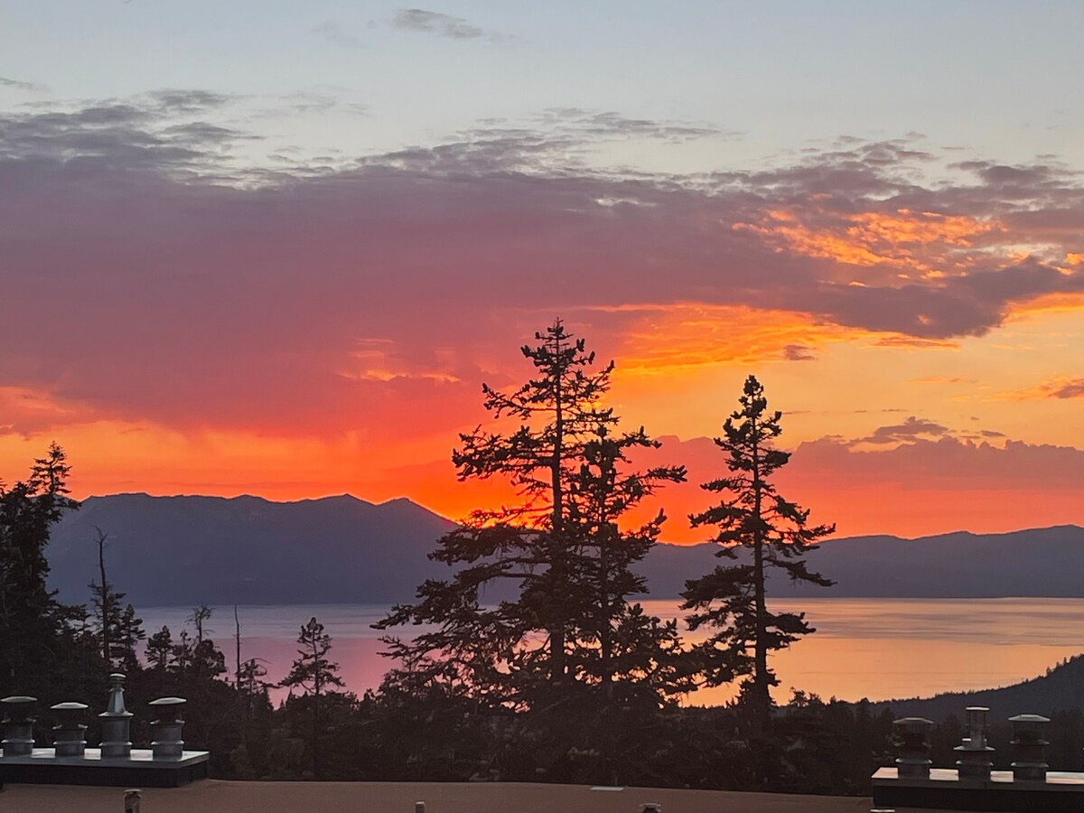 A vibrant sunset is captured over the lake, with layers of orange, pink, and purple hues filling the sky. Silhouetted pine trees frame the view, enhancing the natural beauty of the landscape. The tranquil water reflects the colorful sky, creating a serene atmosphere.
