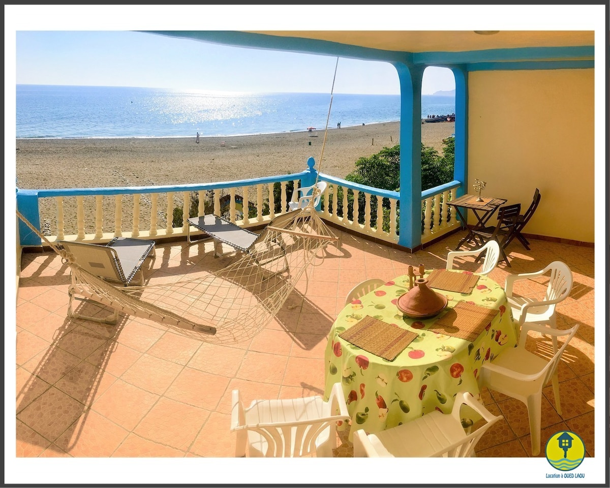 A spacious terrace offers a scenic view of the beach and Mediterranean Sea. The area is furnished with a round table and chairs, complemented by a hammock, creating an inviting space for relaxation. Sunlight reflects off the water, enhancing the serene setting.