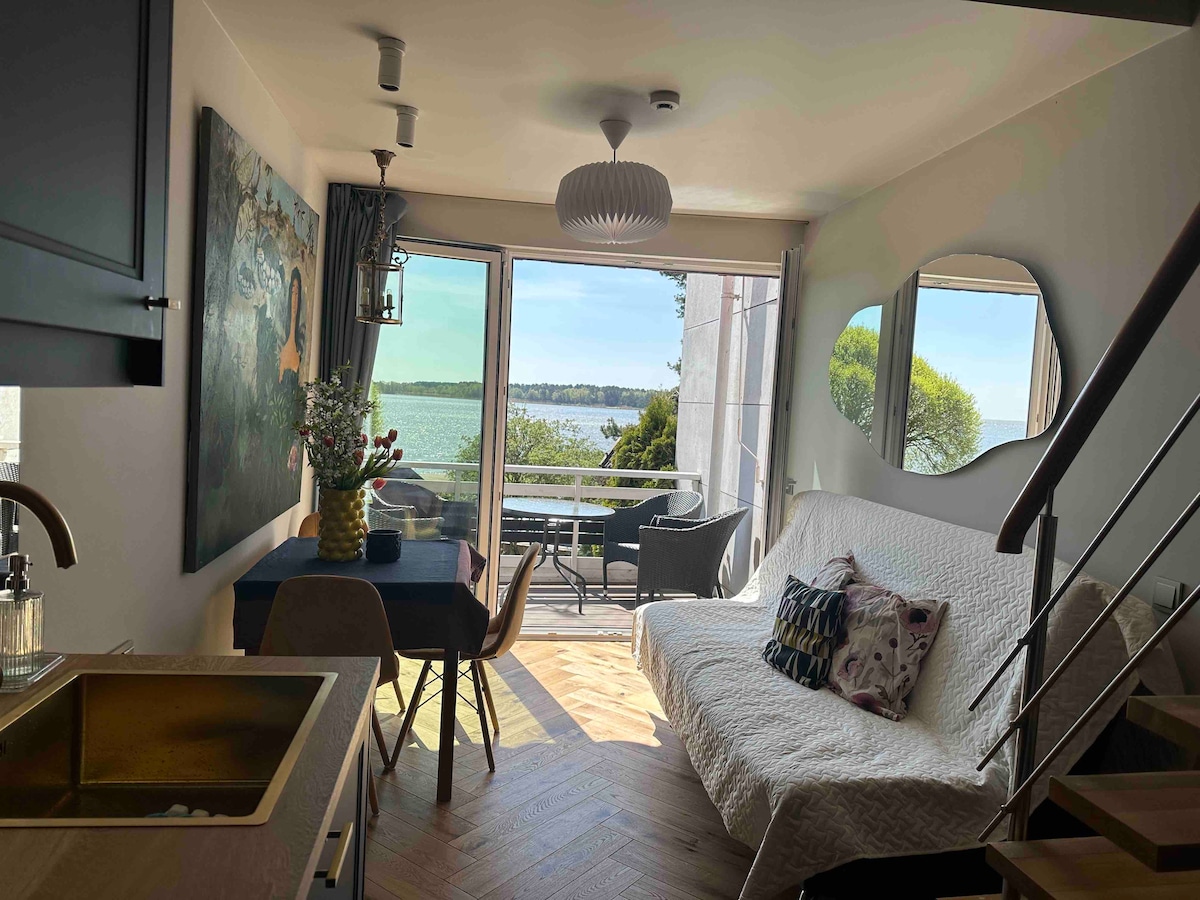 A bright living area features a cozy sofa bed and a dining table set against a large window, offering a view of the water. The space is complemented by a colorful wall art piece and a round mirror, creating a sense of openness.