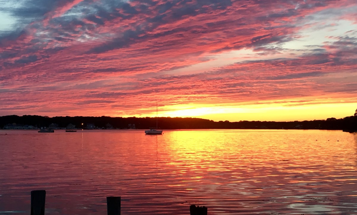 A serene view of the bay at sunset, with vibrant colors reflecting on the water's surface. Silhouettes of trees and a calm horizon are visible, alongside a sailboat anchored gently. The sky showcases a blend of warm oranges, pinks, and purples, creating a peaceful atmosphere.