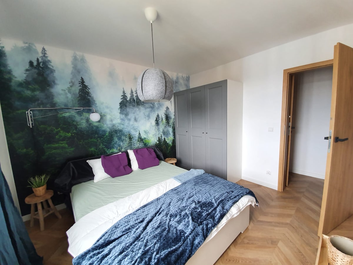 A tranquil bedroom features a spacious bed with pillows in white and purple, complemented by a soft blue blanket. A nature-themed mural depicting a forest enhances the room. A large wardrobe stands to one side, and a door leads to the additional hallway.