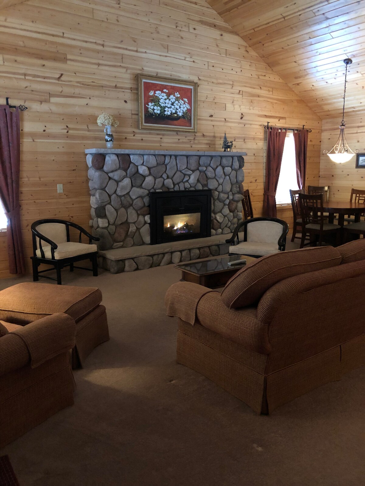A spacious great room features a large stone fireplace as the focal point, surrounded by comfortable seating. Natural wood walls enhance the cabin feel, while windows with rich curtains allow soft light to enter. A dining area with wooden chairs and a table is visible in the background.