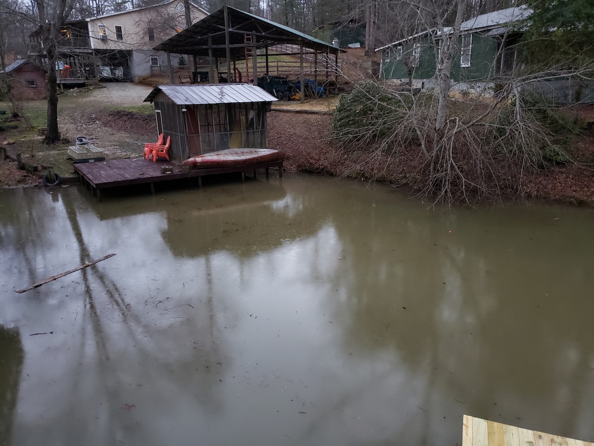 A calm pond reflects the surrounding trees and structures. A small covered dock is visible, accompanied by a pair of red chairs. The area is bordered by dry leaves and bare branches, creating a natural setting.