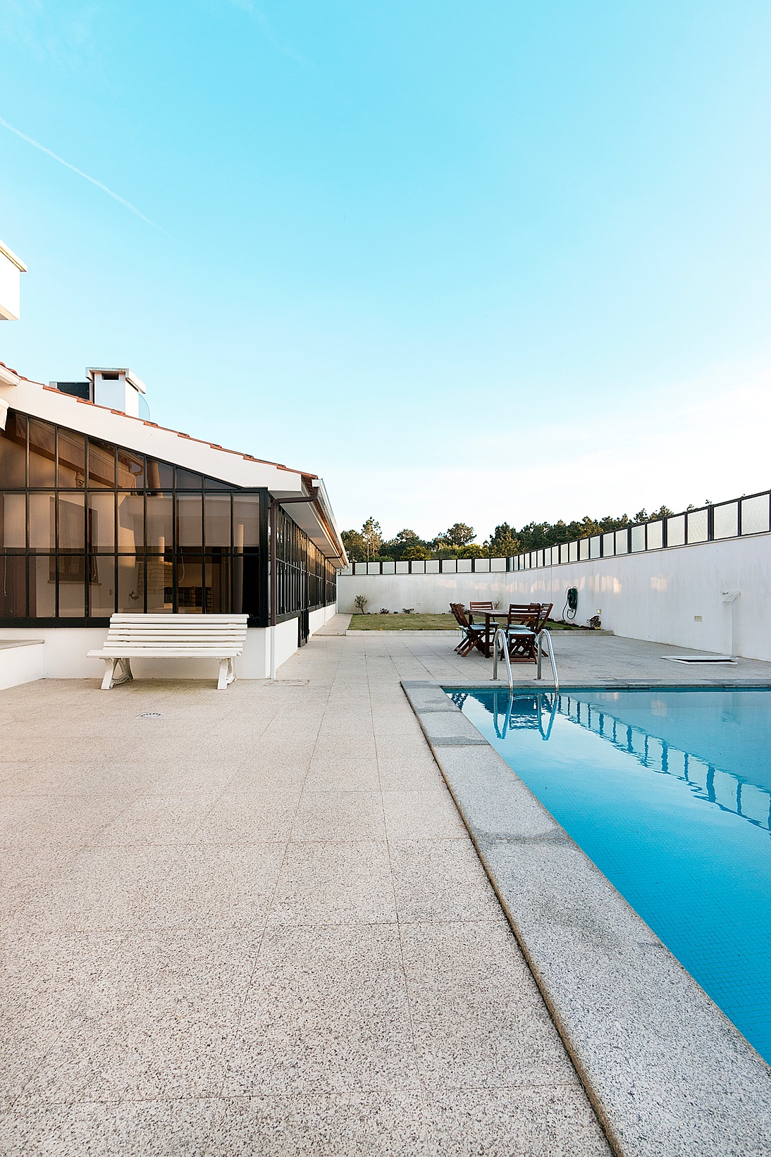A spacious outdoor area is presented, featuring a private pool beside a well-maintained terrace. Sun loungers and seating arrangements are visible, offering opportunities for relaxation and dining. The structure's glass walls enhance the connection between indoor and outdoor settings, with a clear blue sky visible above.