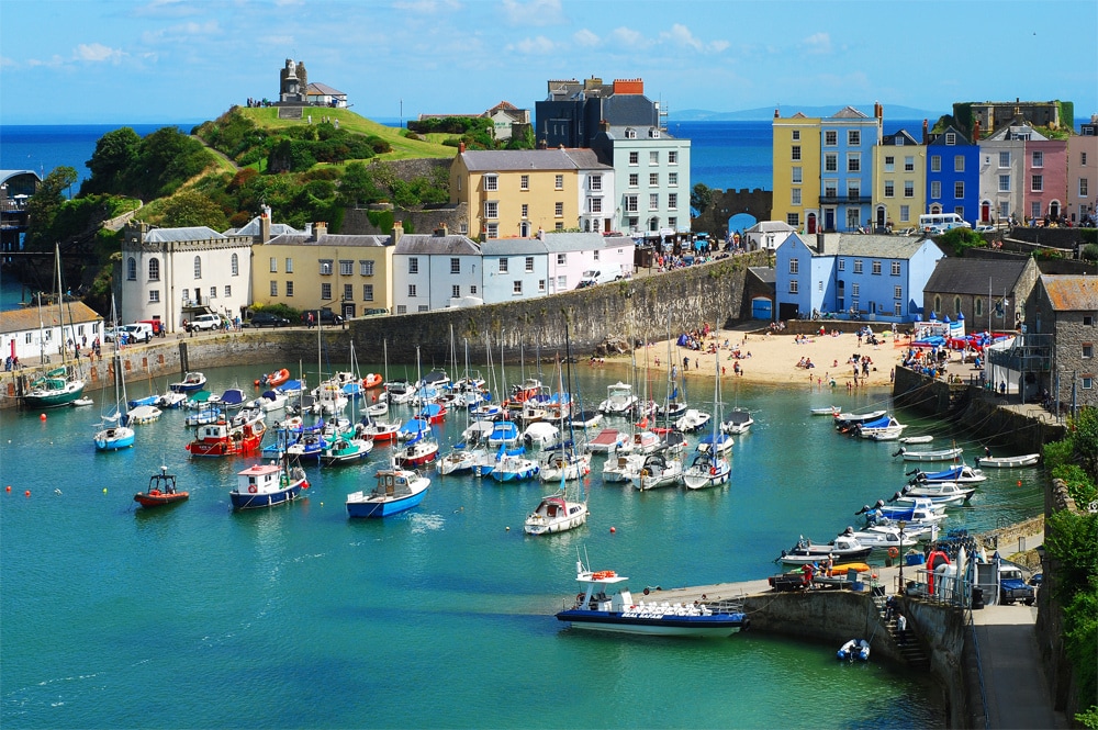 A scenic view of a vibrant harbor, featuring a variety of boats anchored in clear waters. The shoreline reveals a sandy beach with people enjoying the sun, while colorful houses line the waterfront, creating a lively atmosphere against a backdrop of lush green hills.