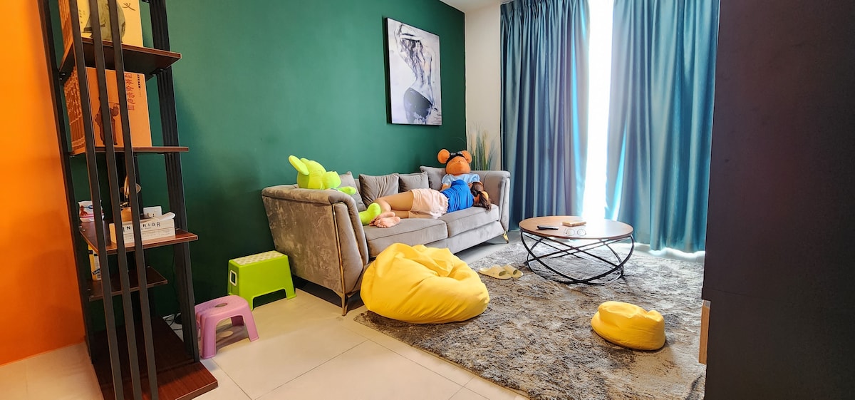 A comfortable living area features a gray sofa adorned with playful stuffed animals. A round coffee table sits center stage atop a soft rug. Colorful bean bags and a small green stool enhance the welcoming space, while large windows invite ample natural light.