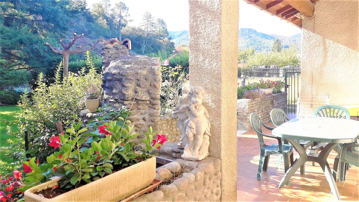 A serene outdoor area features a stone sculpture alongside vibrant flowers in a planter. A dining table and chairs are visible on a patio, with a garden and lush trees in the background. The mountains can be seen in the distance under a clear sky.