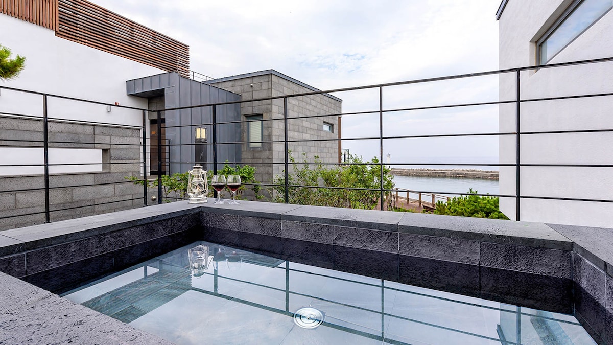 A private outdoor hot tub is surrounded by a sleek stone deck. Glass railings provide an unobstructed view of the coastline and the nearby sea. A minimalist architectural design characterizes the modern structure behind, complemented by the greenery of surrounding plants.