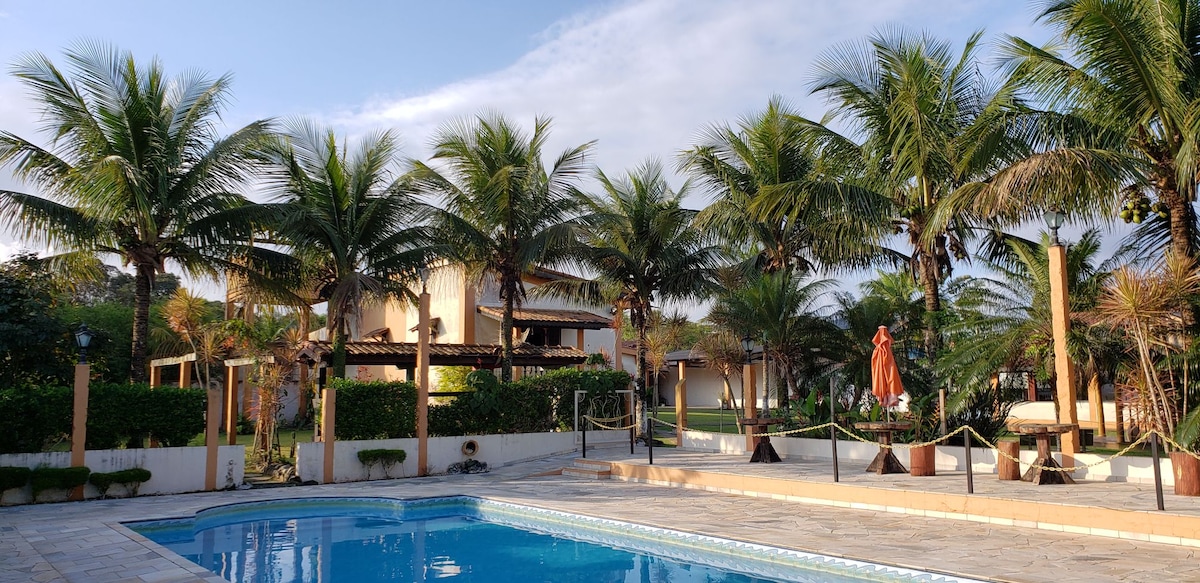 A scenic outdoor area showcases a refreshing swimming pool bordered by palm trees. The pool area is framed by lush greenery, with comfortable seating options and a gazebo in the background. A clear blue sky contributes to the inviting atmosphere, emphasizing relaxation and leisure.