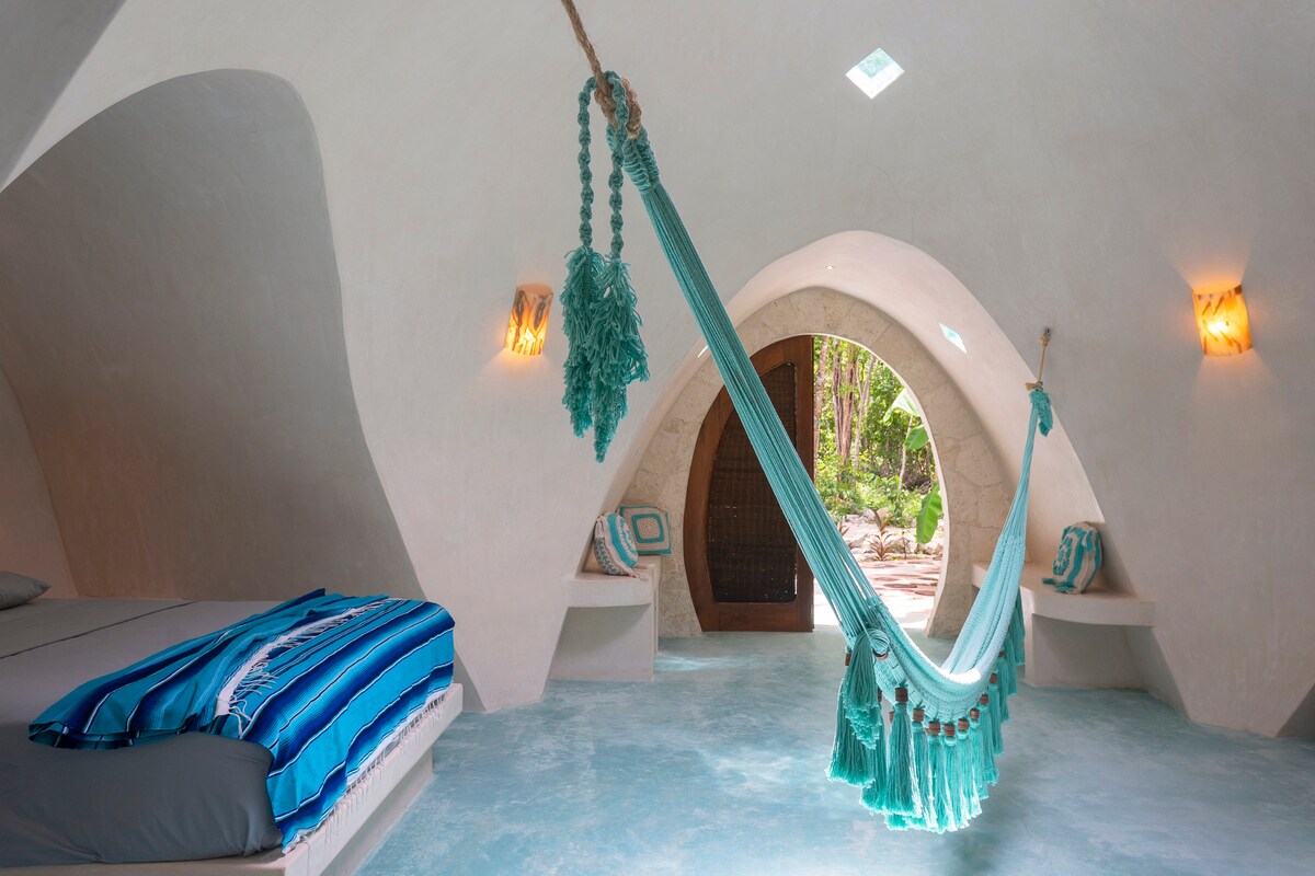 A cozy interior of a Mayan dome features curved white walls and a spacious bed adorned with blue and gray linens. A turquoise hammock is suspended nearby, complemented by decorative pillows. Natural light filters through small windows, illuminating the serene surroundings of the jungle.
