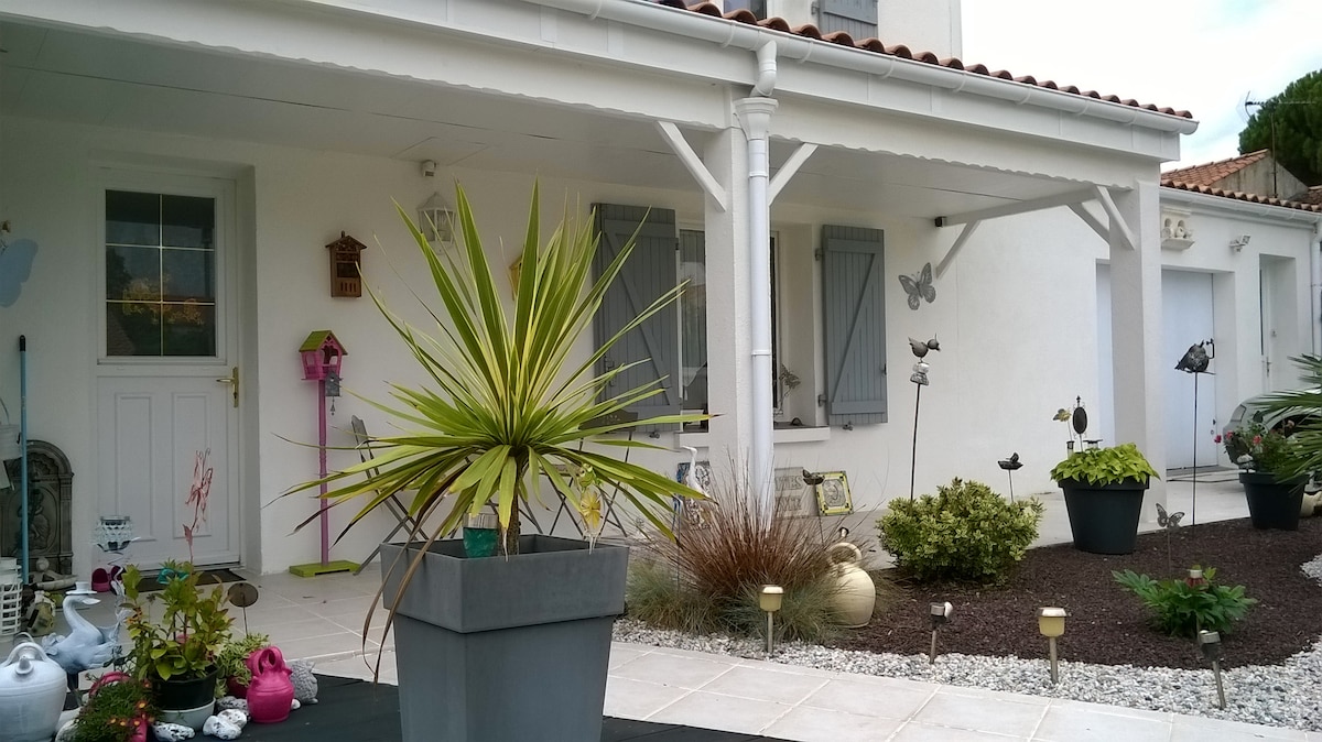 An exterior view of a light-colored house, showcasing a covered porch and decorative elements. Potted plants are arranged prominently, including a large palm and smaller flowers. The entrance is framed by closed shutters and several garden ornaments, creating a serene and welcoming ambiance.