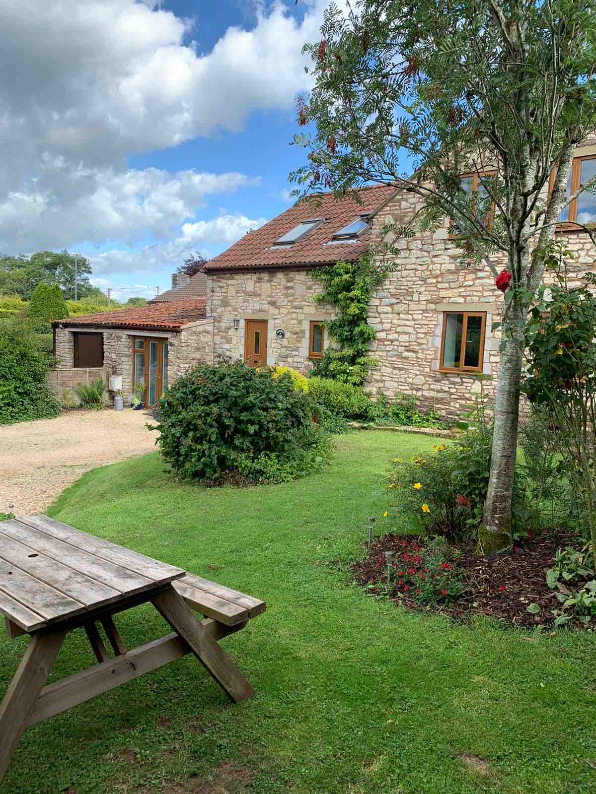 A charming stone building is surrounded by a well-maintained garden featuring grassy areas, flowering plants, and trees. A picnic table is positioned on the lawn, offering a space for outdoor relaxation. Clear skies and soft clouds create a serene backdrop.