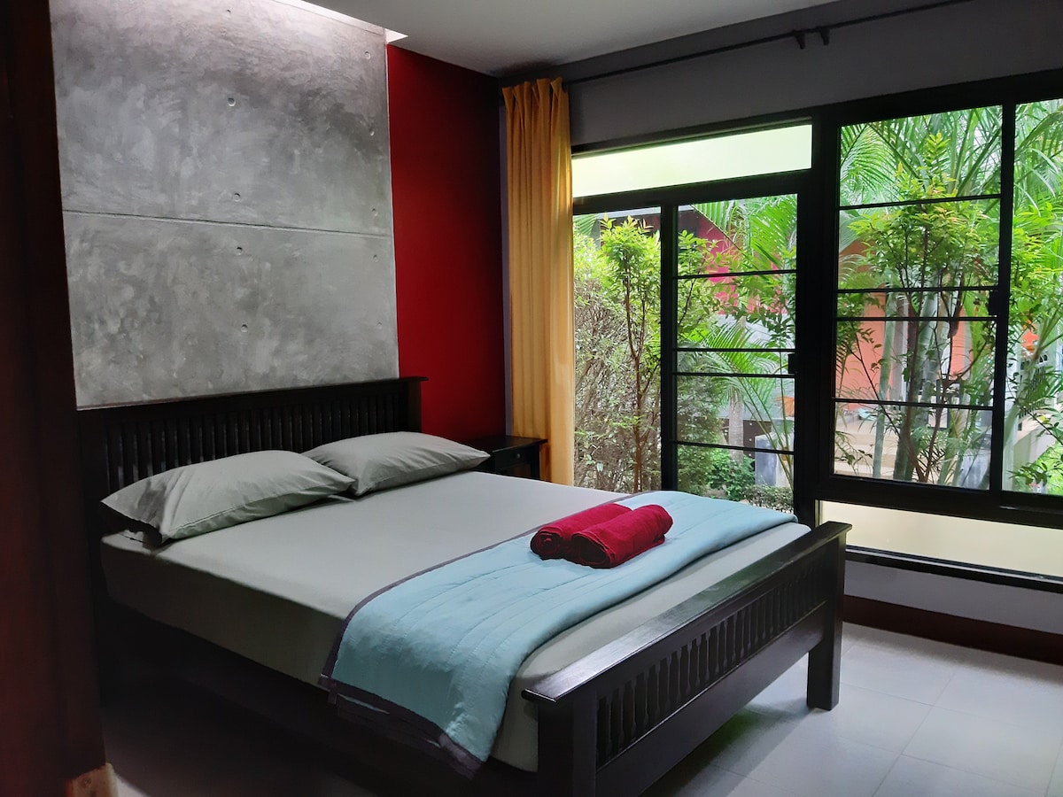 A modern bedroom is presented with a spacious bed topped with neutral bedding and a folded towel. Large windows allow natural light to fill the room, revealing views of surrounding greenery, while a feature wall of textured concrete adds an interesting design element.