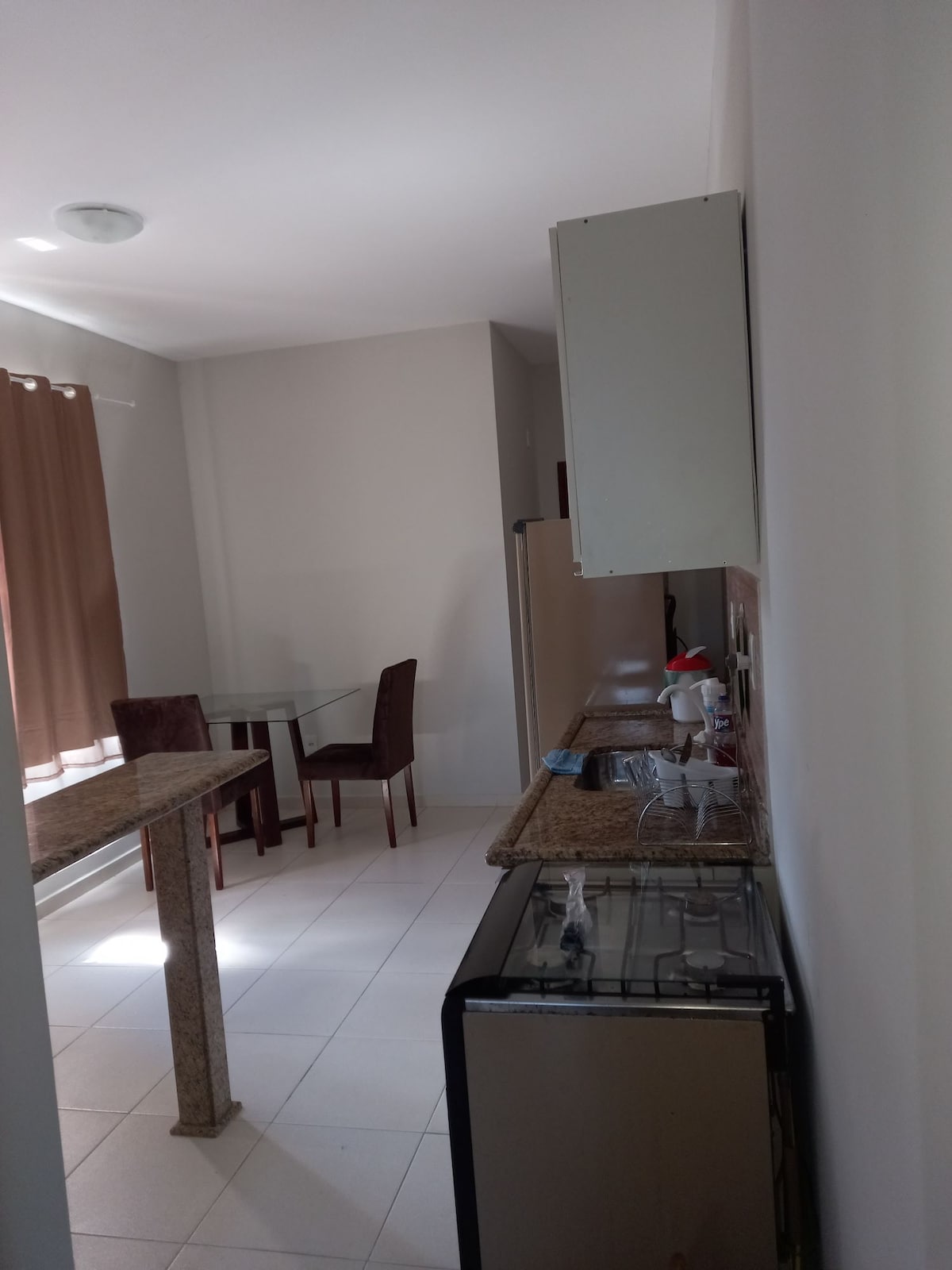 A modern kitchen area features a granite countertop and a sleek stove. A compact dining table with four chairs is positioned near the window, which is adorned with light brown curtains. Soft natural light fills the space, highlighting the clean, tiled floor.