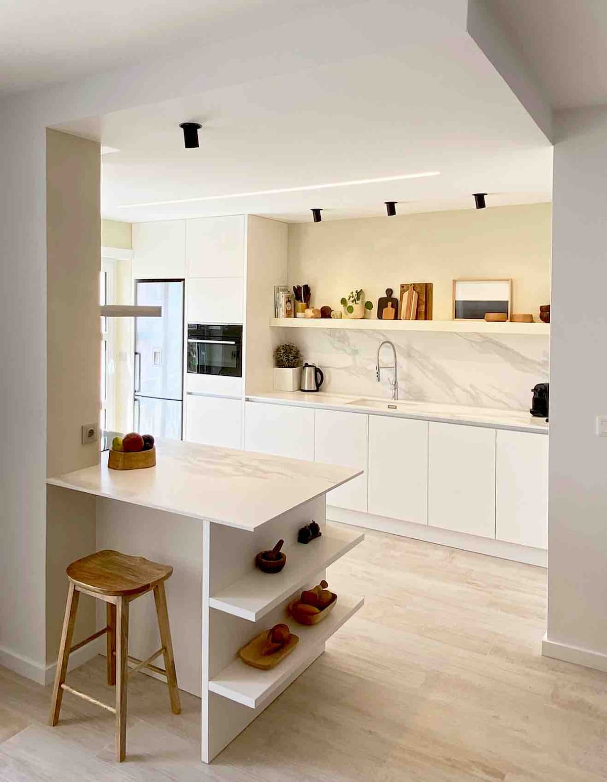 A modern kitchen is shown with a white countertop and sleek cabinetry. Subtle lighting highlights the space, and a wooden stool is positioned beside a minimalist island. Kitchen essentials, including a kettle and a coffee maker, are neatly arranged on the counter.