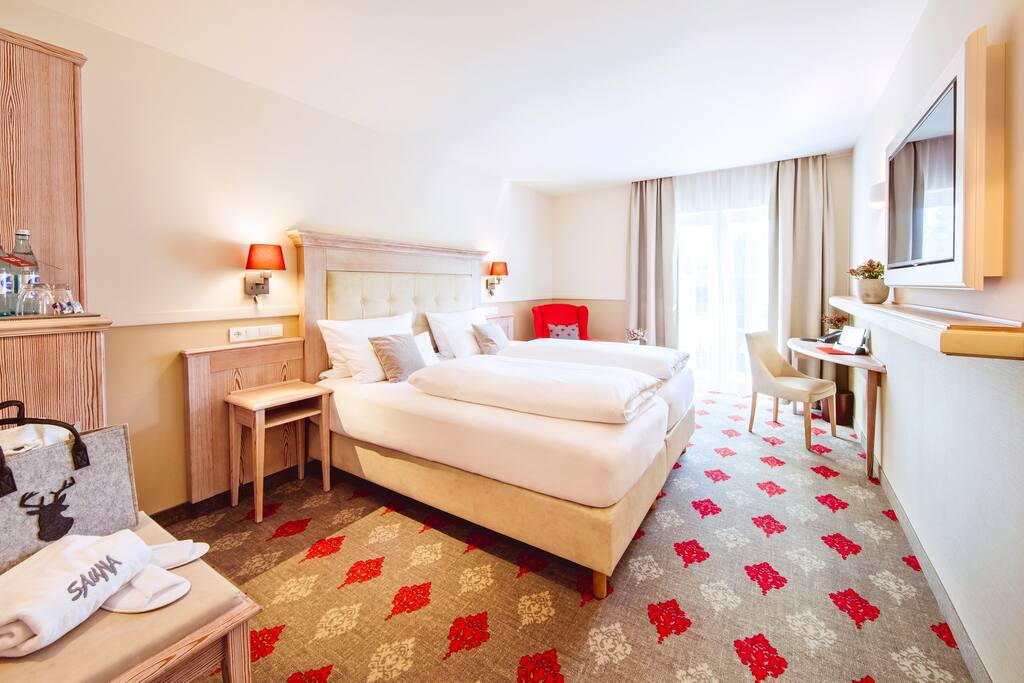 A modern room features a spacious bed with light-colored bedding set against neutral walls. A side table and a small desk are positioned near a window with light curtains. Decorative elements include a patterned carpet in red and beige tones.