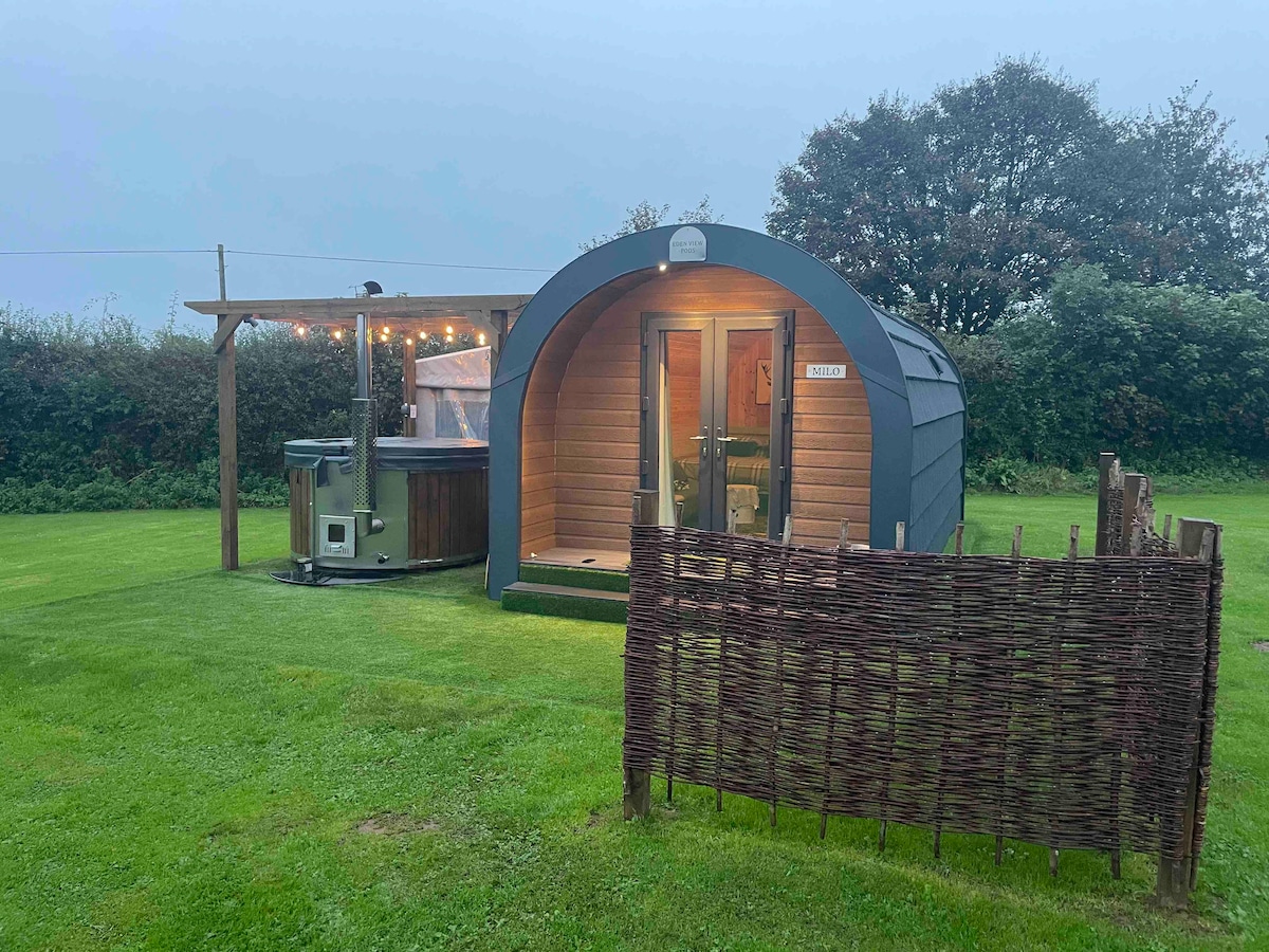 A glamping pod is viewed from the garden, set against a green landscape. The pod features a rounded roof and wooden paneling. A wood-fired hot tub is positioned nearby under a sheltered area adorned with string lights, while a rustic fence adds an element of privacy.