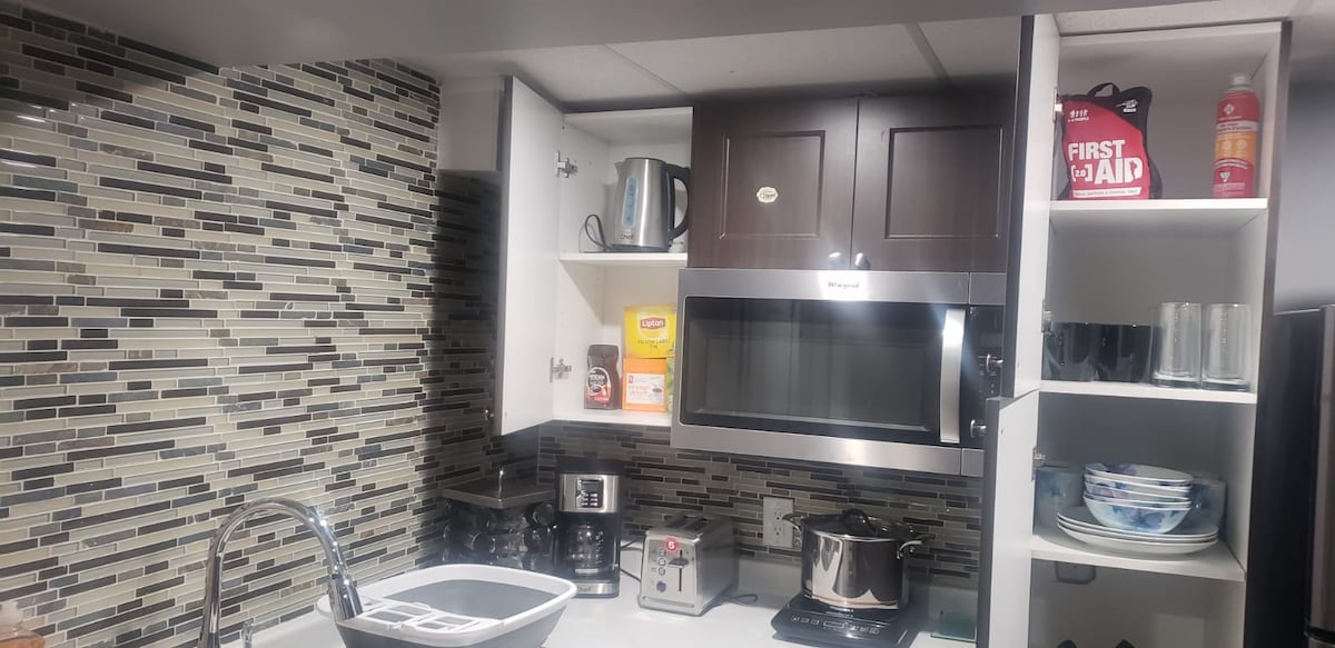 A functional kitchenette is displayed with modern cabinetry and a tiled backsplash. Essential appliances include a kettle, microwave, and coffee brewer. A variety of cookware and cutlery are organized neatly on shelves, with a sink at the forefront for convenience and ease of use.