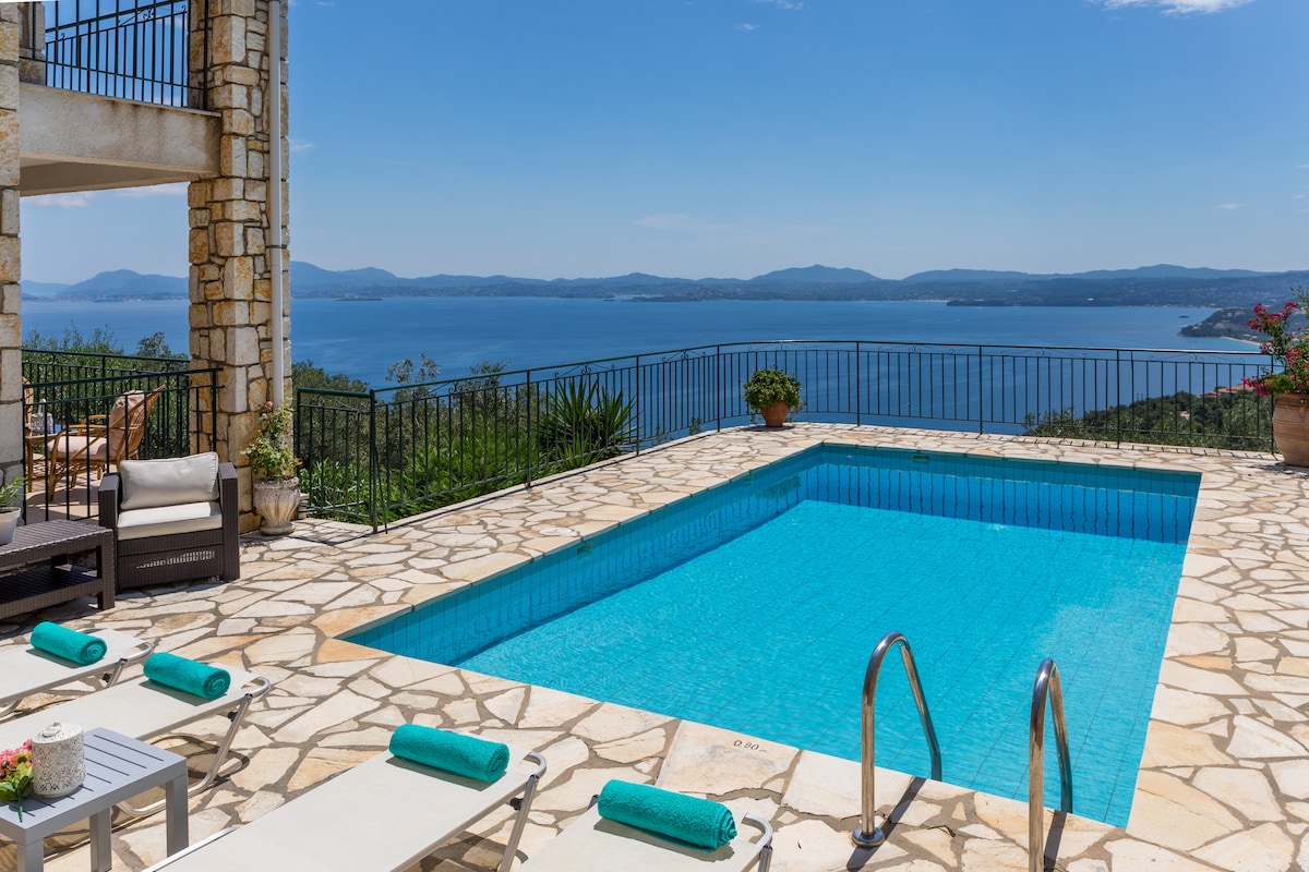 A private pool is set within a stone terrace, framed by a panoramic view of the distant coastline and mountains. Lounge chairs with rolled towels line one edge of the pool, while planters with greenery enhance the natural setting.