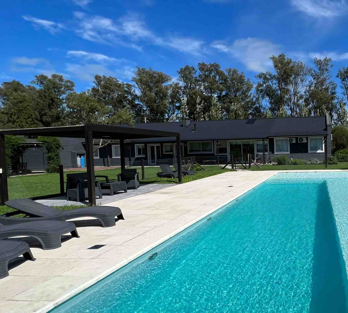 A spacious outdoor area features a crystal-clear swimming pool bordered by a sun deck. Comfortable lounge chairs are arranged around the pool, and the backdrop includes a single-story house with large windows, surrounded by lush greenery and mature trees.