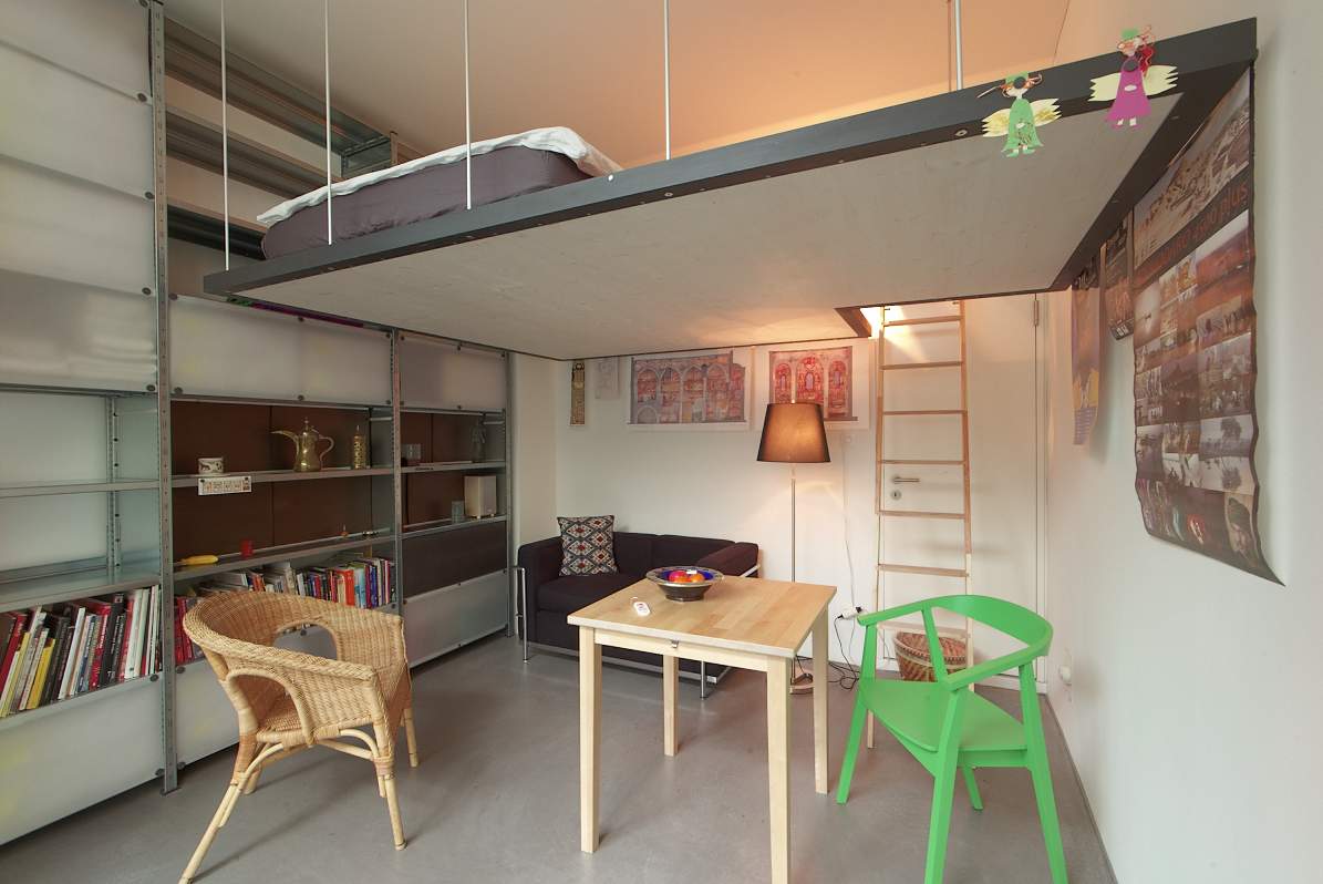 A compact studio features a sleeping gallery above the living area, accessible by a ladder. A cozy seating area includes a sofa and a small dining table, surrounded by shelves filled with books. Light-colored walls and a mix of furniture create a functional space.