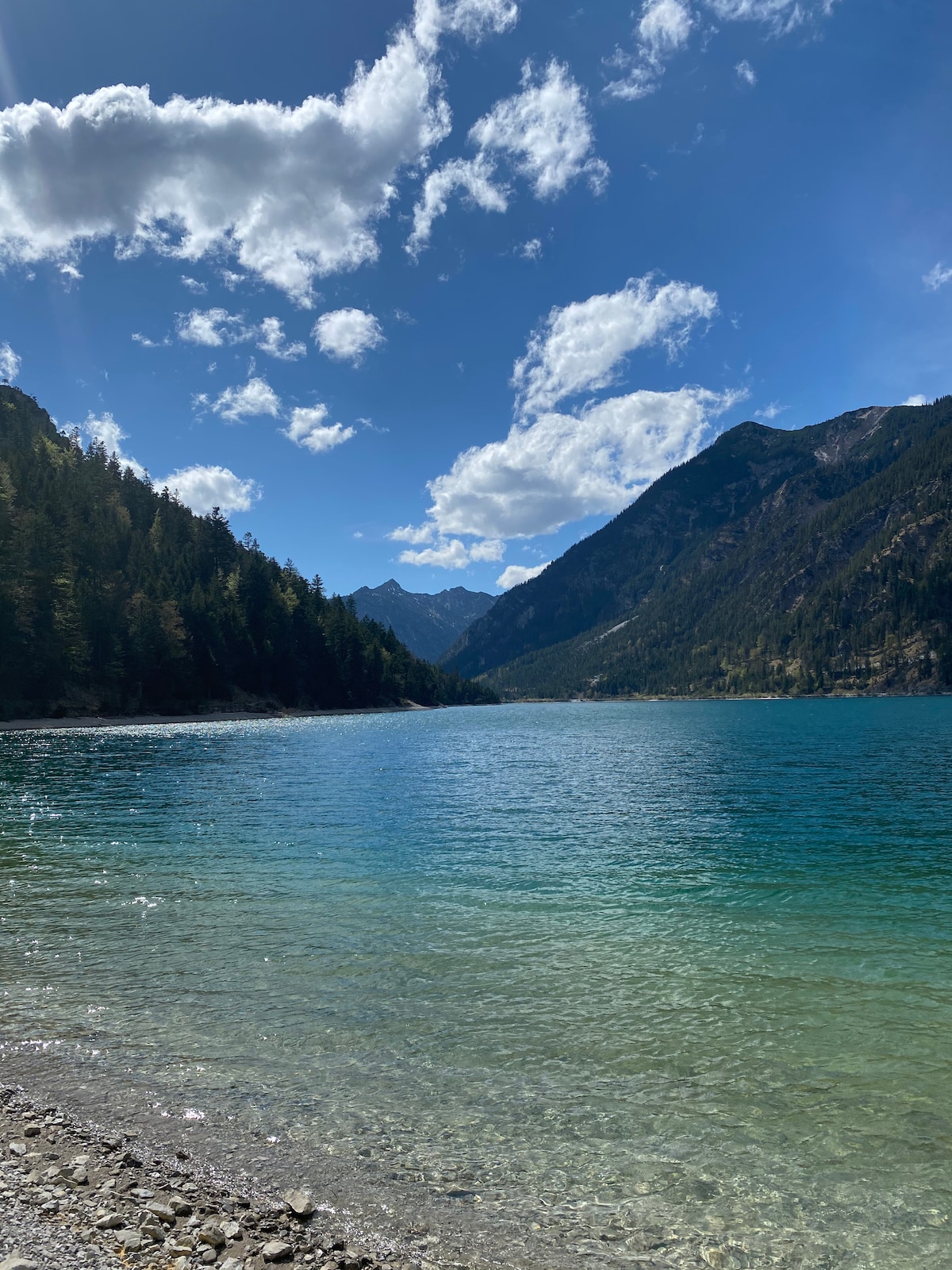 A serene landscape features a vast body of water reflecting the clear blue sky, surrounded by towering mountains and lush greenery. Soft clouds drift above, contributing to a calm atmosphere. The shoreline is marked by smooth pebbles leading to the crystal-clear water.
