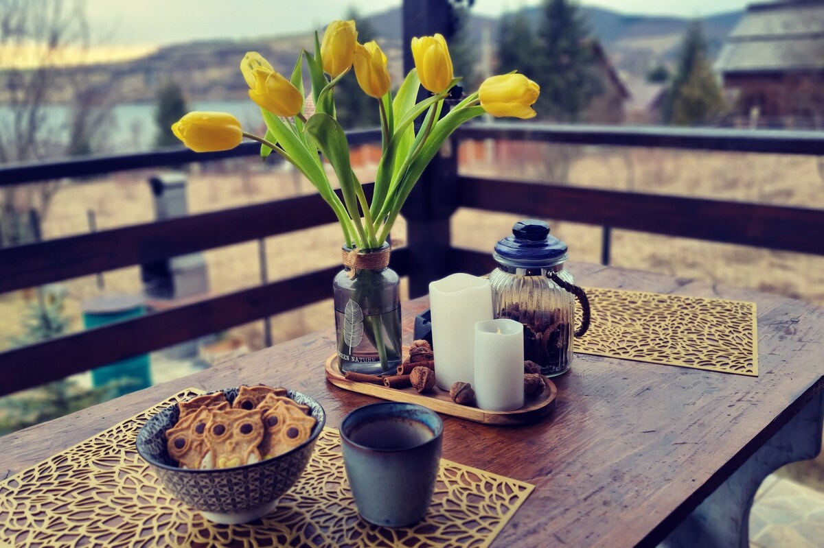A wooden table is set for relaxation, featuring a vase of yellow tulips, a bowl of cookies, a cup, and candles arranged on a decorative tray. The scene is framed by a view of the tranquil landscape and distant lake.