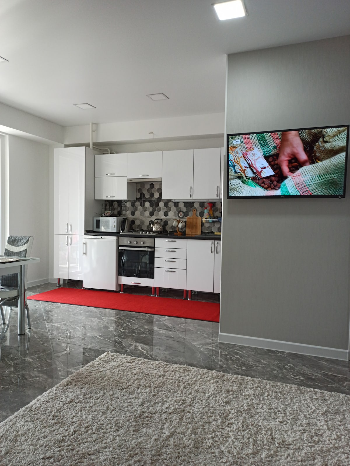A modern kitchen is depicted with sleek white cabinetry and stainless steel appliances. A gray backsplash with a geometric pattern complements the space. A dining area with transparent chairs is visible, along with a rug that adds texture to the flooring. A television is mounted on the wall.
