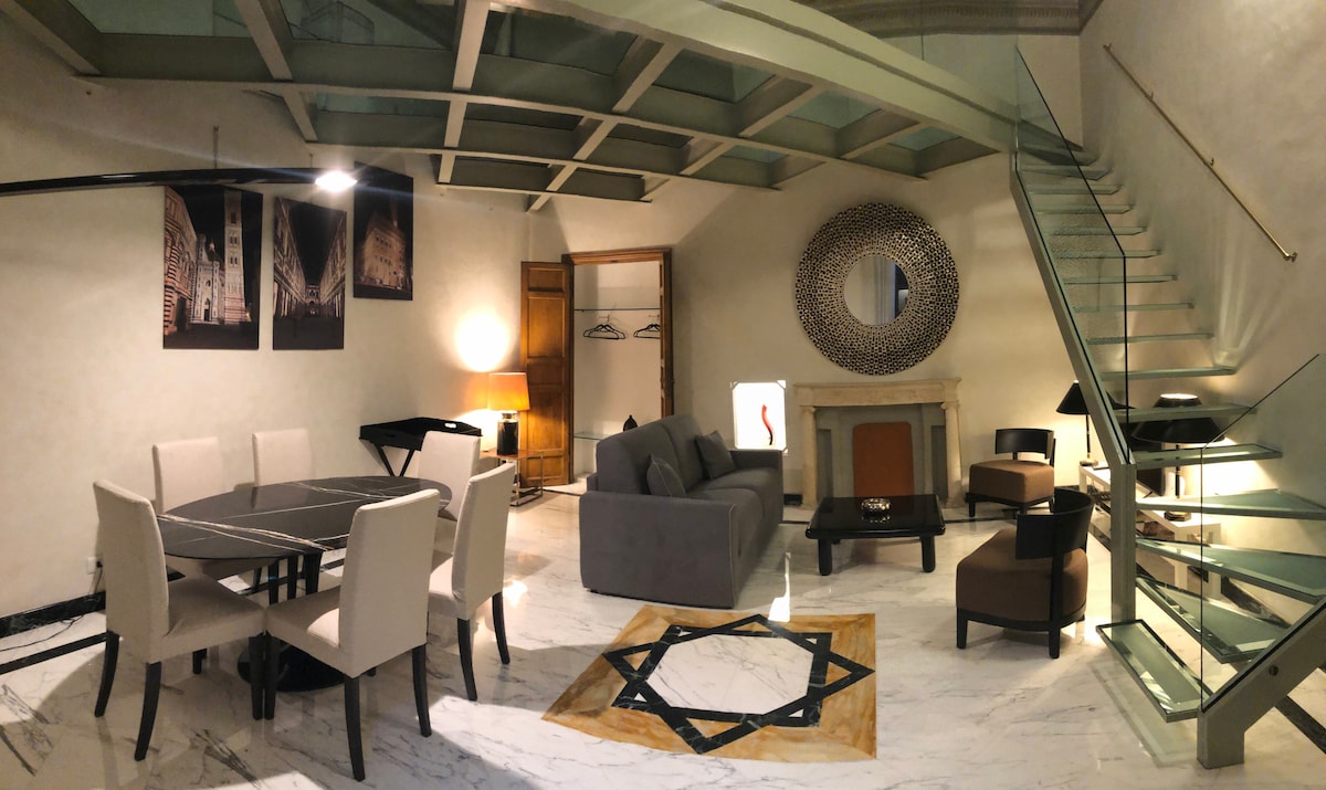 The living area features a spacious layout with a glass loft above, a modern dining table with six chairs, and a comfortable seating arrangement. Artworks adorn the walls, and the marble flooring is complemented by an accent rug. A decorative mirror is positioned above the fireplace.