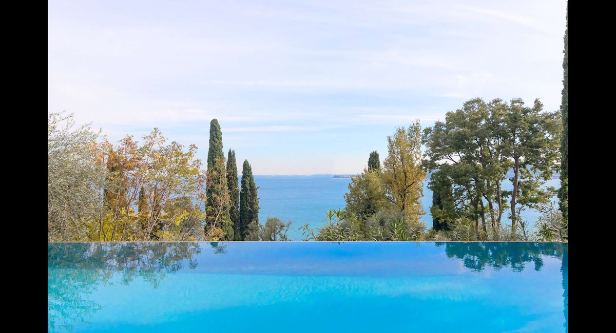 An infinity pool extends towards the lake, creating a seamless connection with the water. Surrounding tall trees and colorful foliage frame the view of the tranquil lake and distant horizon, offering a peaceful natural setting. The sky above is bright with soft clouds.
