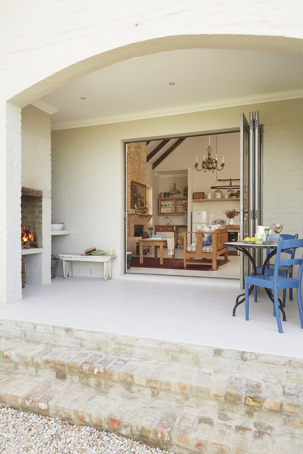 The outdoor patio features a dining table with blue chairs, set against a backdrop of a charming indoor kitchen visible through opened stacker doors. A cozy fireplace is positioned to the left, enhancing the inviting atmosphere of the space.