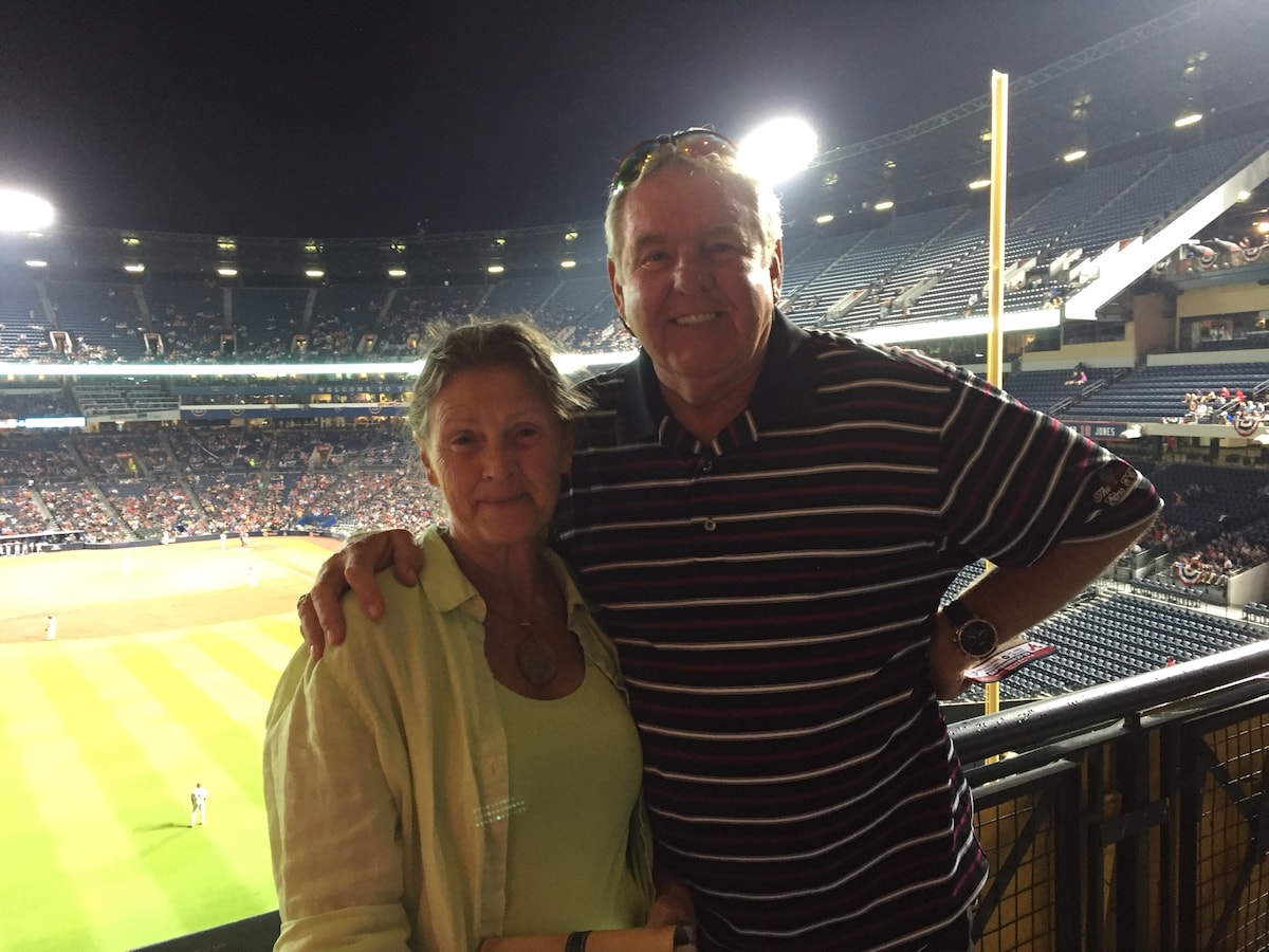 A couple stands together at a sports venue, with a view of the stadium filled with spectators in the background. Bright lights illuminate the venue, creating a vibrant atmosphere. The couple is positioned against a railing, with the field visible beyond them.