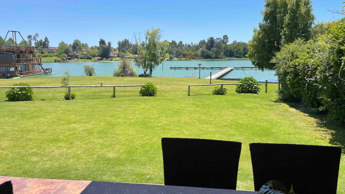 A expansive green lawn extends toward a serene lake, which reflects a clear blue sky. A wooden dock is visible on the water, with trees and shrubs lining the perimeter, offering a natural and inviting landscape.