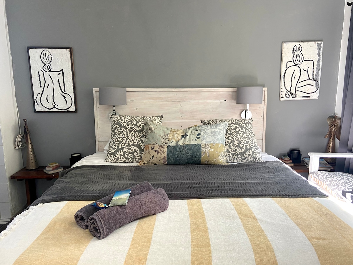 A cozy sleeping area features a queen-sized bed adorned with patterned pillows and a neutral throw blanket. Artwork is displayed on the walls, adding character. Two bedside lamps provide soft lighting, creating a relaxed ambiance.