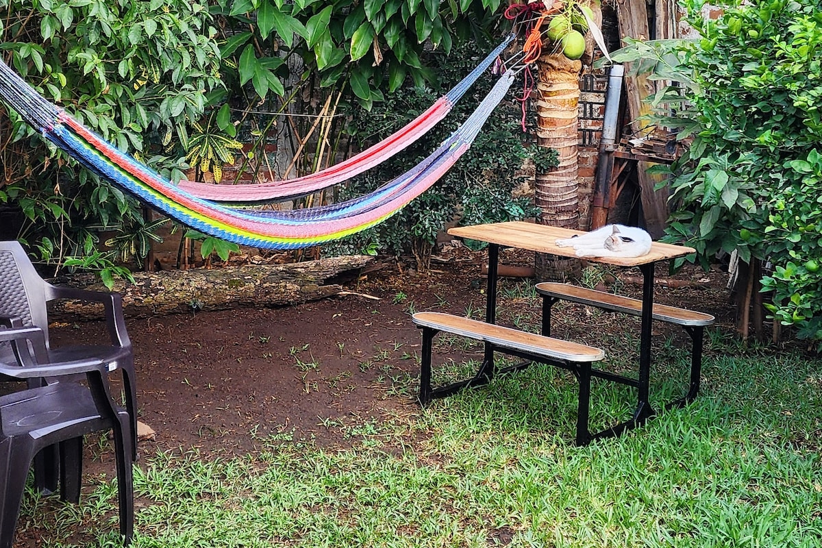 A spacious outdoor patio is depicted, featuring a wooden table with bench seating. A vibrant, multicolored hammock is draped between two trees, surrounded by lush greenery. The ground is covered in grass, creating a relaxing environment for outdoor dining or leisure.