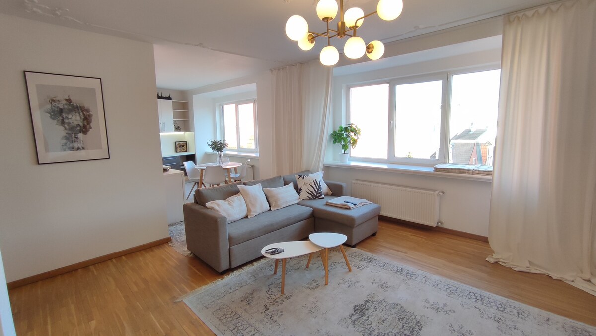 A spacious living area is highlighted by large windows that allow natural light to fill the room. A comfortable gray sofa is paired with a wooden coffee table on a patterned rug, with a small workspace visible in the background near the kitchen.