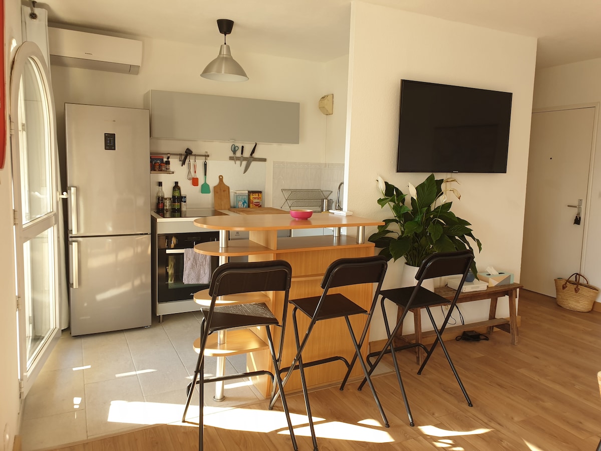 The open kitchen features a modern design with a refrigerator, an oven, and a decorative plant on the countertop. Four black chairs are positioned around a wooden bar. A large flat-screen TV is mounted on the wall, providing an inviting space for relaxation.