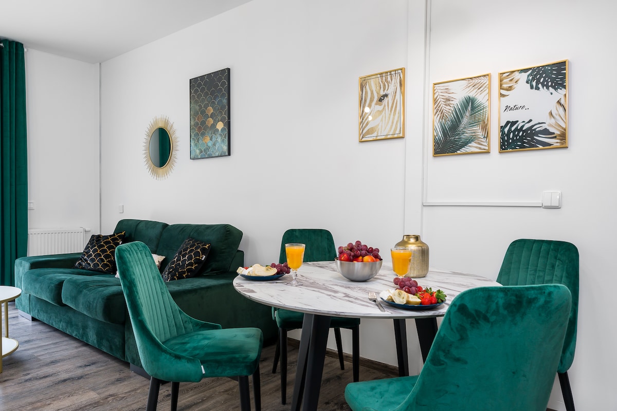 A modern living space is showcased, featuring a round table set for four with colorful drinks and fruit. A comfortable green sofa with decorative pillows enhances the inviting atmosphere, while framed artwork adorns the walls, contributing to the overall aesthetic.