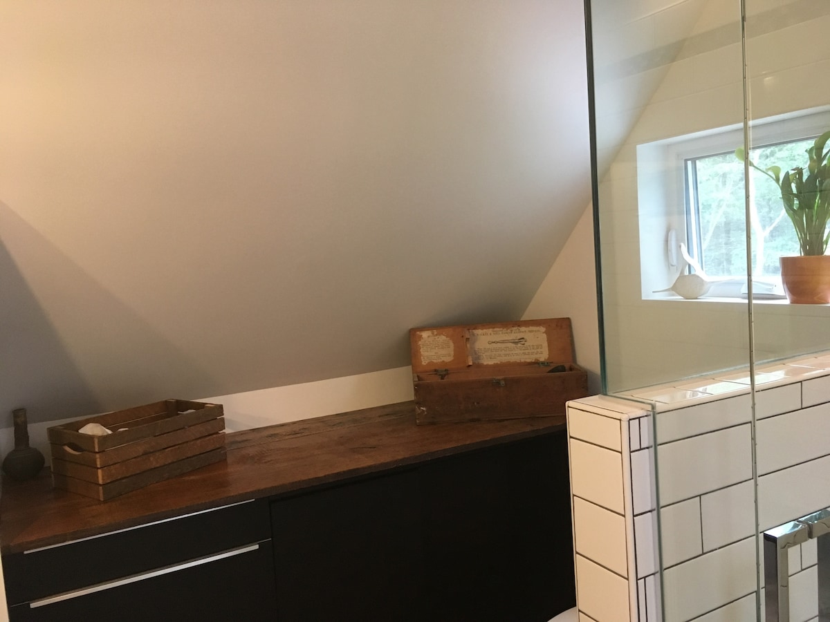 A bathroom corner showcases a wooden counter under a sloped ceiling, complemented by a decorative wooden box and a small potted plant on the windowsill. The adjacent glass shower reflects light, providing an open feel, while the clean lines of the tiled wall enhance the modern aesthetic.