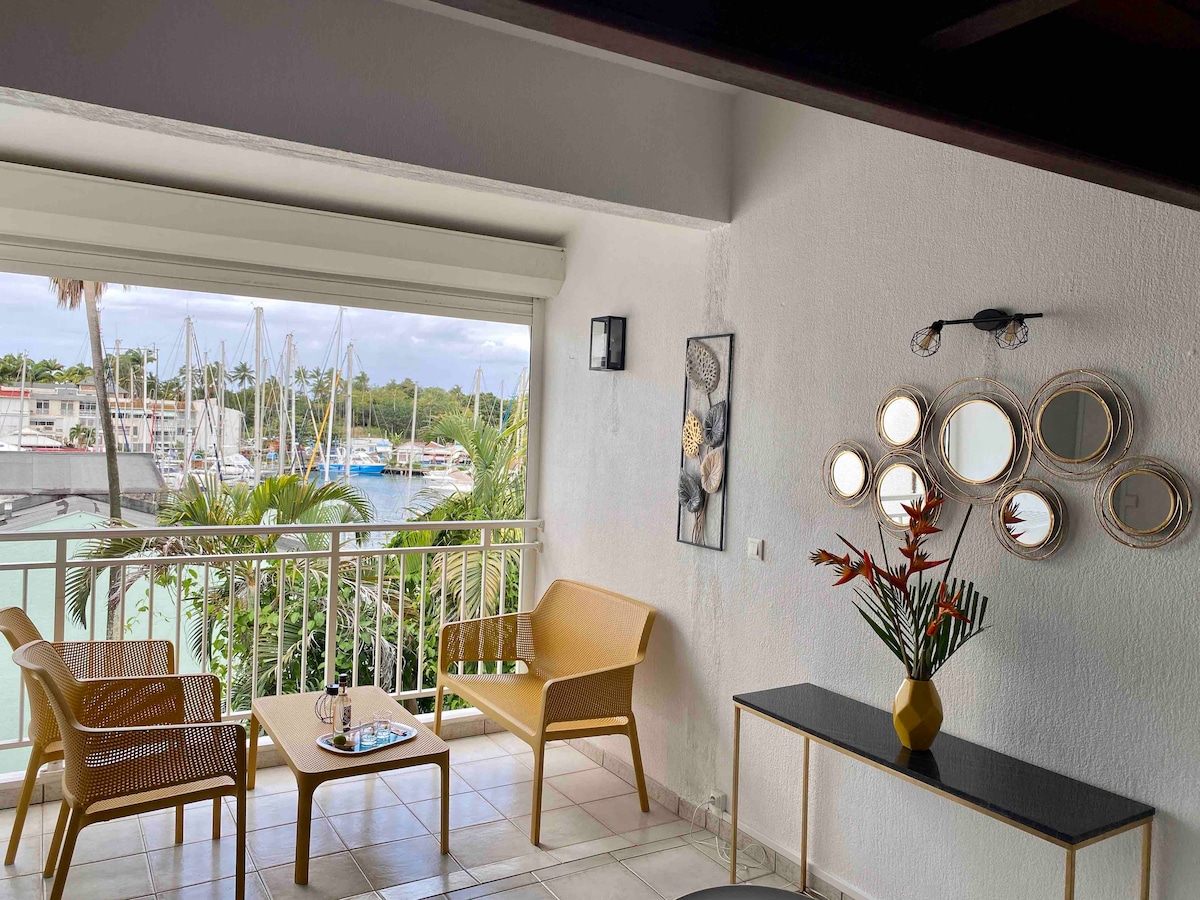 A cozy seating area features two yellow chairs and a small table, positioned to offer a view of the marina through large sliding glass doors. Tropical plants and artwork enhance the space, while a sleek side table adds a modern touch.