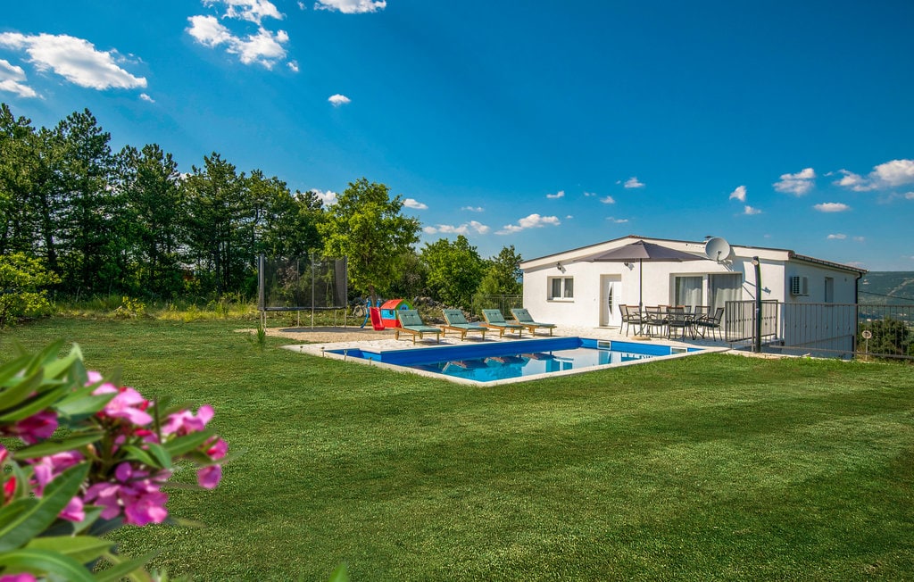 An inviting outdoor area features a private pool surrounded by lounge chairs, with lush green grass extending in all directions. The house is visible in the background, complemented by a clear blue sky dotted with clouds.