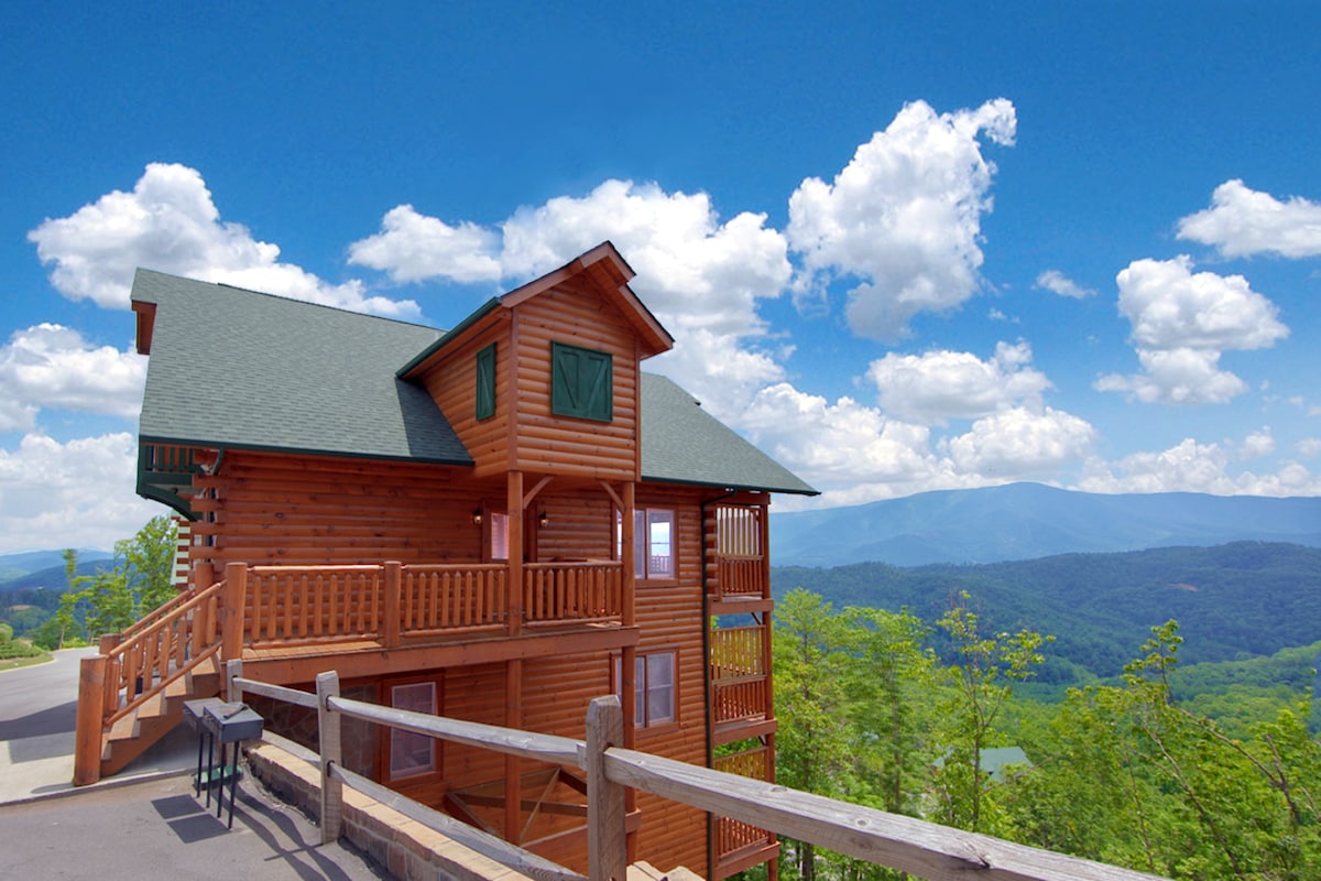 A spacious log cabin is presented against a backdrop of rolling mountains and a bright blue sky dotted with white clouds. The well-maintained exterior includes multiple balconies and a distinctive green roof. A railing surrounds the deck, enhancing the panoramic views.