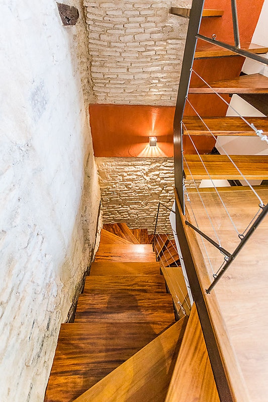 A spiral wooden staircase is visible, leading down within a brick and stone interior. The warm lighting accentuates the curves of the railing and the wooden steps, creating a seamless transition between the different levels of the home.