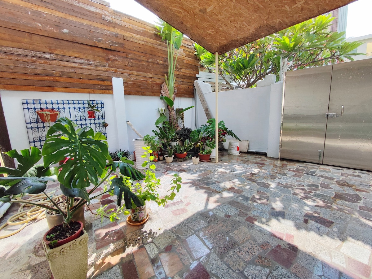 A sheltered outdoor area is filled with various potted plants, creating a relaxed atmosphere. The stone flooring provides a natural feel, while a wooden structure offers shade. A metal storage cabinet is visible along one side, complementing the greenery.