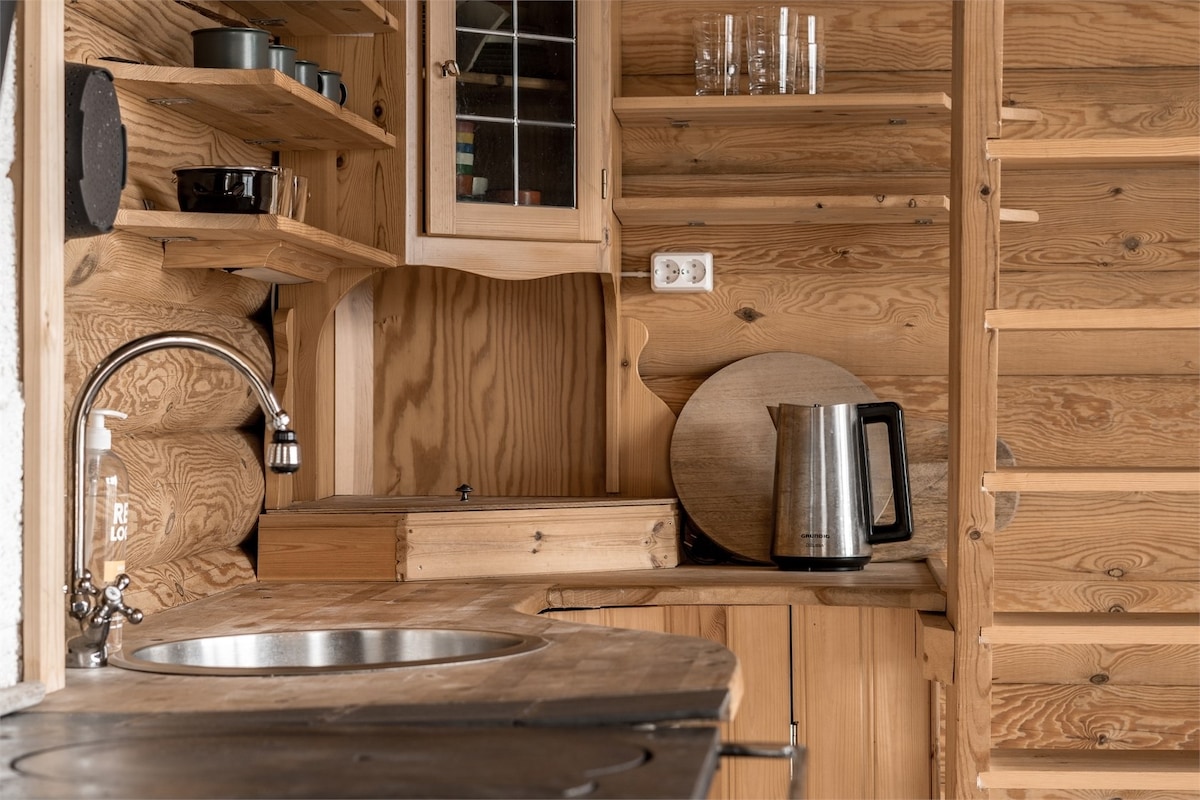 A compact kitchenette features wooden cabinetry and shelves, showcasing a kettle, utensils, and dishware. A stainless-steel sink is integrated into a wooden countertop. Natural light highlights the warm tones of the log walls and the staircase leading to the loft.