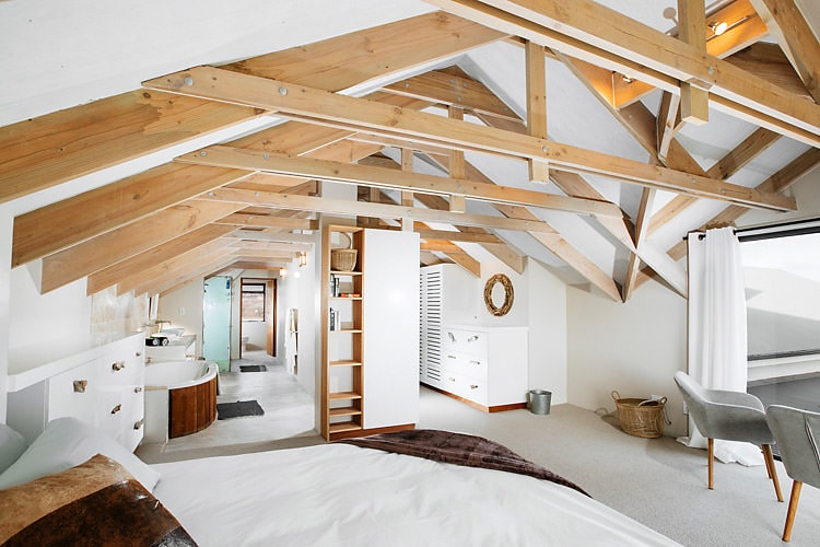 A spacious bedroom is highlighted by a high, wooden-beamed ceiling and large windows. A comfortable bed is positioned in the foreground, with light fabrics creating a calming feel. The room features a cozy seating area and offers access to an en-suite bathroom, enhancing the overall comfort and functionality.