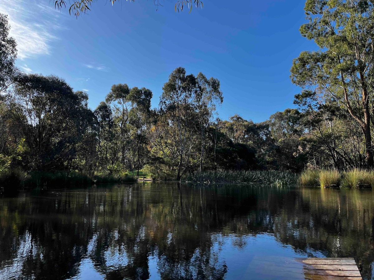 A calm water body reflects the blue sky and surrounding greenery. Lush trees and shrubs frame the scene, creating a serene atmosphere. The stillness of the water enhances the natural beauty, inviting a sense of peace and tranquility.
