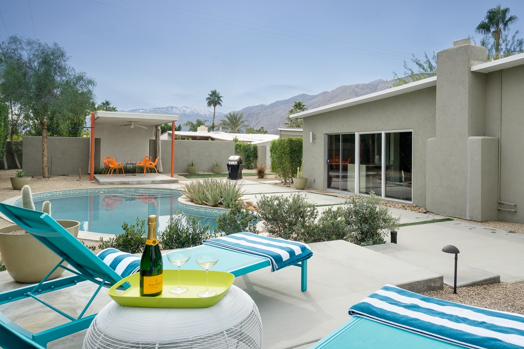 A serene outdoor space is highlighted by a refreshing pool and desert mountain views. Poolside lounge chairs are arranged beside a small table featuring champagne and glasses. Lush greenery and minimalist decor complement the contemporary setting, with a covered patio visible in the background.